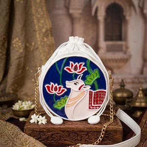 May include: White, round handbag with a blue center featuring a cow and lotus flower design. The bag has a white drawstring closure, a white strap, and a gold chain. The bag is displayed on a wooden box.