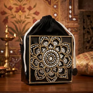 May include: Black square handbag with a floral design in gold and white. The bag has a drawstring closure and a white cord. The background features an ornate wall design and a candle.