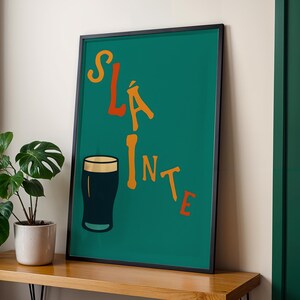 May include: A framed art print with a teal background and the word "Sl&aacute;inte" in orange and yellow lettering. A black pint glass is featured below the text. The print is displayed on a wooden surface next to a potted plant.
