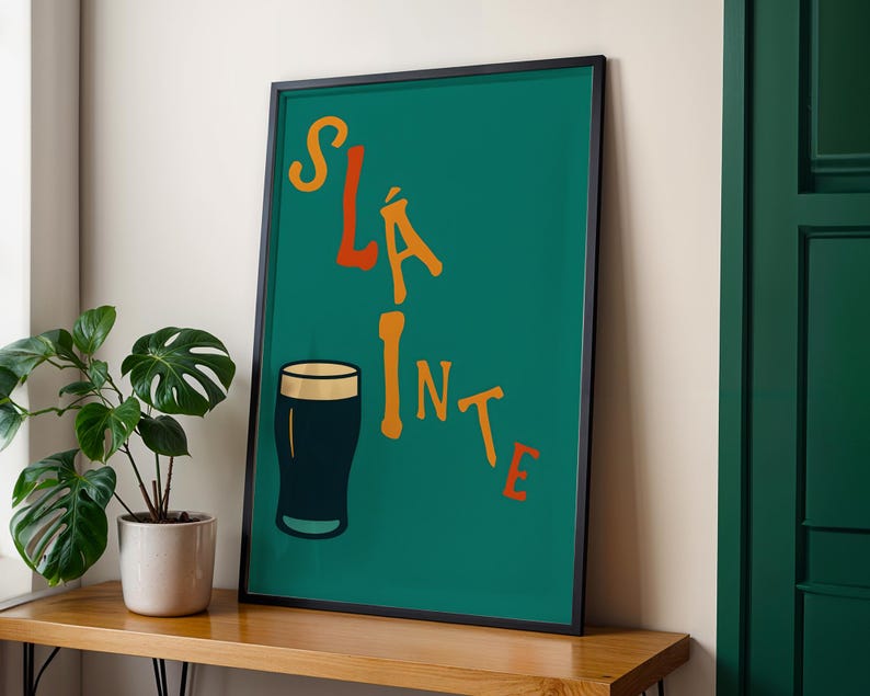 May include: A framed art print with a teal background and the word "Sl&aacute;inte" in orange and yellow lettering. A black pint glass is featured below the text. The print is displayed on a wooden surface next to a potted plant.