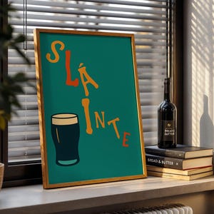 May include: A framed teal print with the word "Sl&aacute;inte" in orange letters and a black pint glass illustration. A bottle of wine and a stack of books are next to the print on a windowsill.