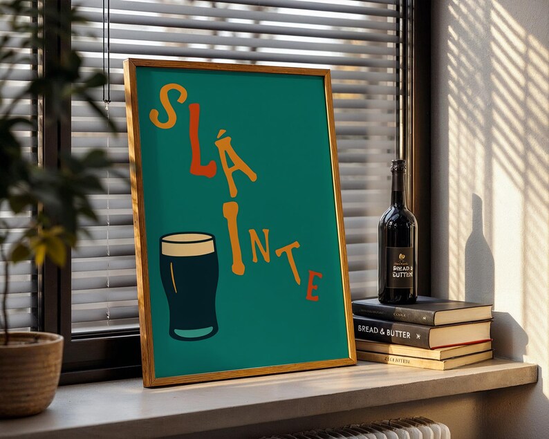 May include: A framed teal print with the word "Sl&aacute;inte" in orange letters and a black pint glass illustration. A bottle of wine and a stack of books are next to the print on a windowsill.