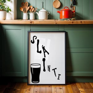 May include: Black framed art print with the word "Sl&aacute;inte" and a pint glass illustration. The black text is in a stylized font. The print is displayed in a kitchen with green cabinets and wooden countertops.