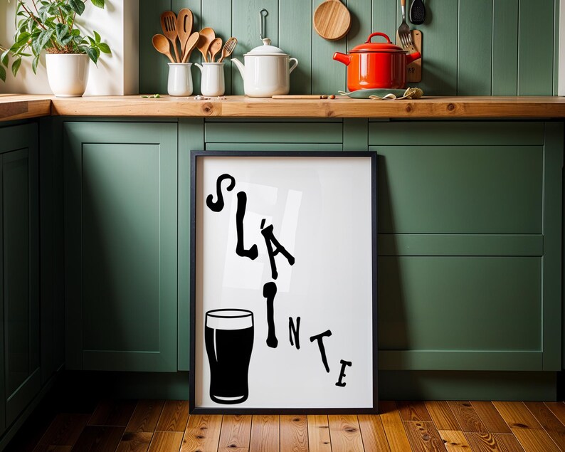 May include: Black framed art print with the word "Sl&aacute;inte" and a pint glass illustration. The black text is in a stylized font. The print is displayed in a kitchen with green cabinets and wooden countertops.