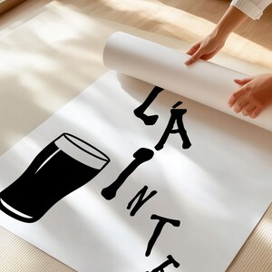 May include: A white poster with a black graphic of a pint glass and the words "LA PINTE" in black. The poster is being unrolled by a person. The poster is on a light-colored rug.