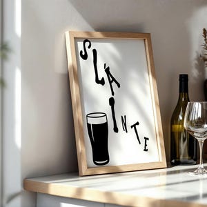 May include: A framed print with the word "Sl&aacute;inte" and a pint glass illustration in black. A wine bottle and glass are next to the frame on a white countertop. The frame has a light wood finish.