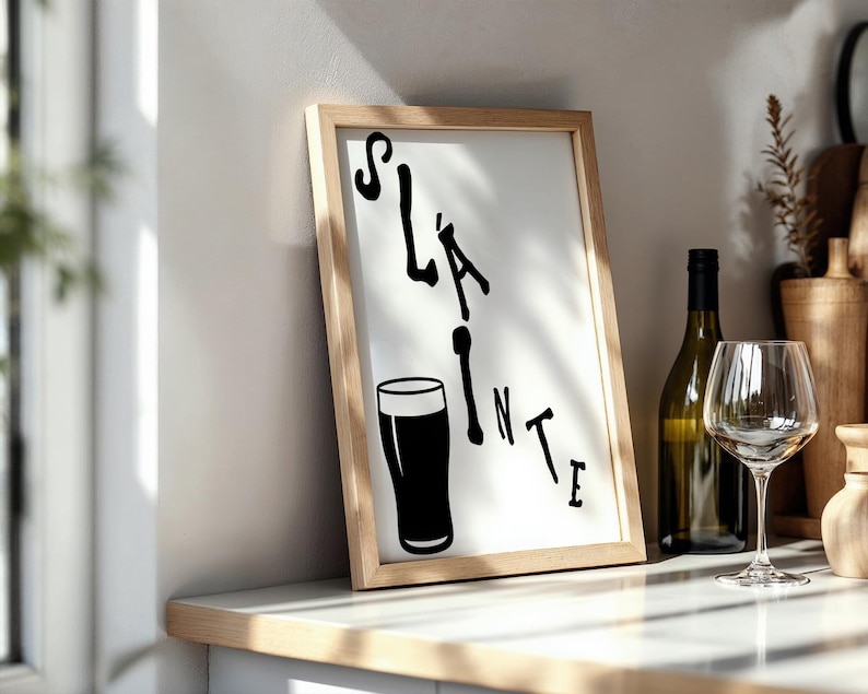 May include: A framed print with the word "Sl&aacute;inte" and a pint glass illustration in black. A wine bottle and glass are next to the frame on a white countertop. The frame has a light wood finish.