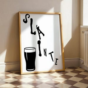 May include: A framed print with the word "Sl&aacute;inte" in black script, accompanied by a black pint glass illustration. The wooden frame surrounds a white background. The print leans against a wall with a checkered floor.