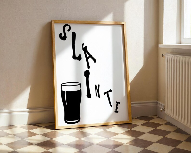 May include: A framed print with the word "Sl&aacute;inte" in black script, accompanied by a black pint glass illustration. The wooden frame surrounds a white background. The print leans against a wall with a checkered floor.