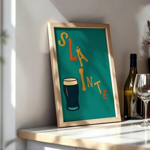 May include: A framed teal print with the word "Sl&aacute;inte" in orange and yellow lettering, along with a dark pint of beer. A wine bottle and glass are visible on a white countertop.