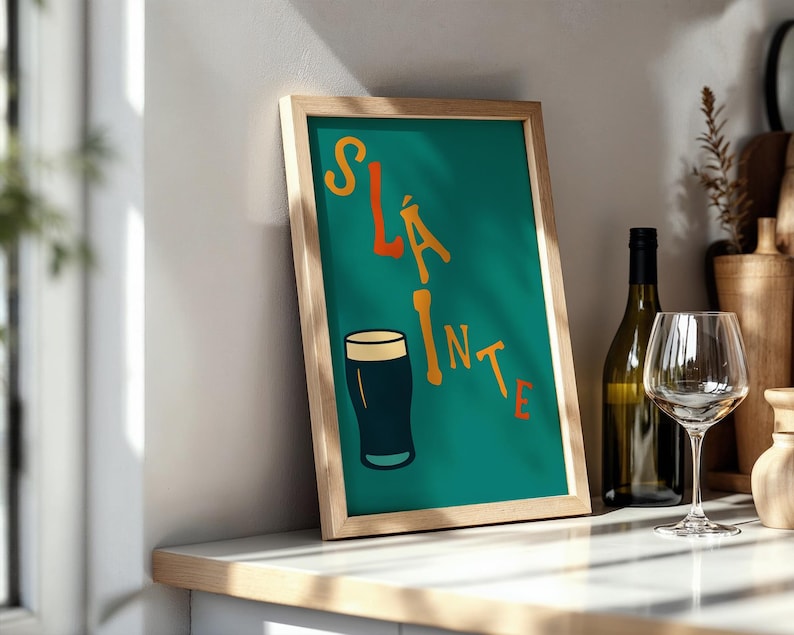 May include: A framed teal print with the word "Sl&aacute;inte" in orange and yellow lettering, along with a dark pint of beer. A wine bottle and glass are visible on a white countertop.