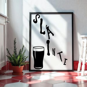 May include: Black framed art print with the word "Sl&aacute;inte" and a pint glass illustration. The artwork is displayed on a white wall, with a small potted plant and a white stool nearby. The floor has a red and white tile pattern.