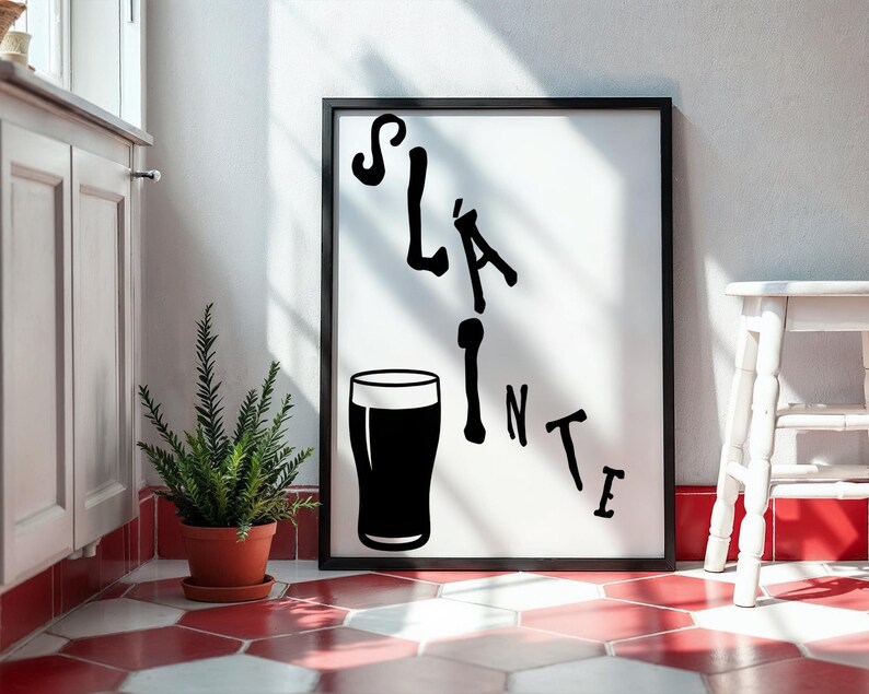 May include: Black framed art print with the word "Sl&aacute;inte" and a pint glass illustration. The artwork is displayed on a white wall, with a small potted plant and a white stool nearby. The floor has a red and white tile pattern.