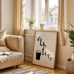 May include: A framed print with the word "Sl&aacute;inte" and a pint glass illustration in black. The artwork is displayed in a bright room with a beige sofa, a window, and a potted plant. The room has a cozy and inviting atmosphere.