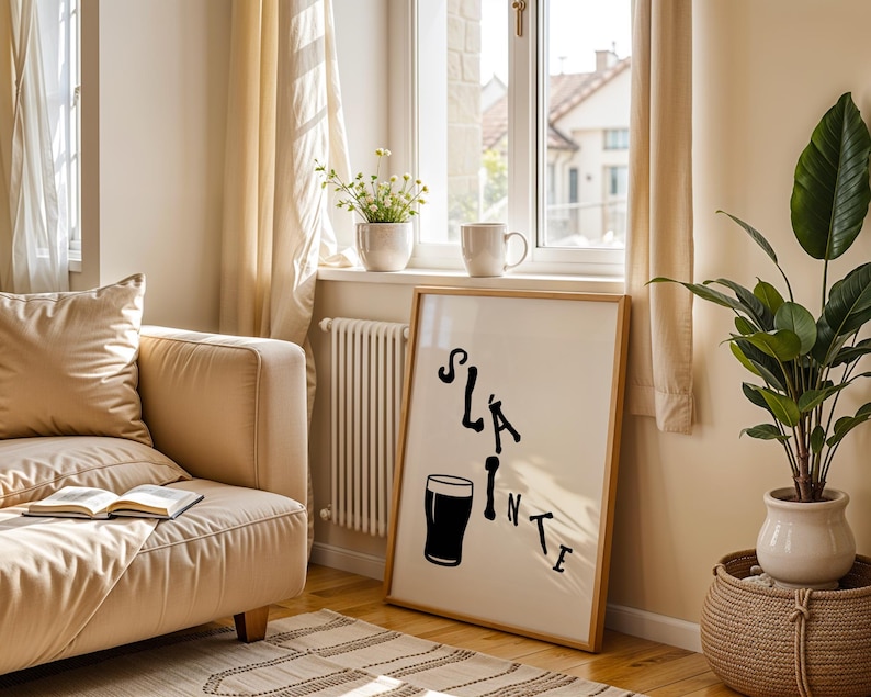 May include: A framed print with the word "Sl&aacute;inte" and a pint glass illustration in black. The artwork is displayed in a bright room with a beige sofa, a window, and a potted plant. The room has a cozy and inviting atmosphere.