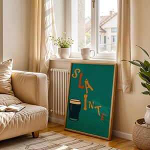 May include: A framed teal poster with the word "SL&Aacute;INTE" in orange and yellow lettering, along with a cartoon pint of stout. The poster is displayed in a living room setting, next to a beige sofa and a potted plant.