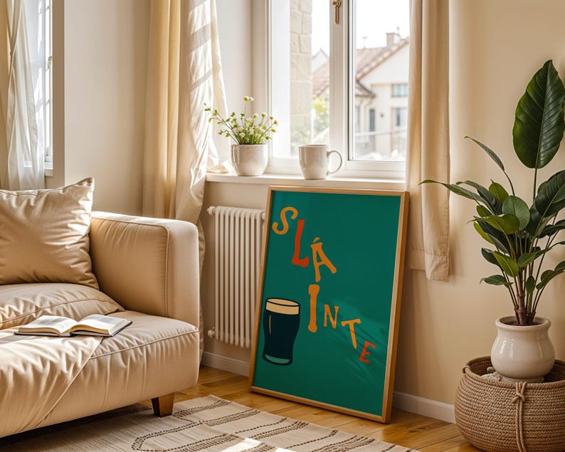 May include: A framed teal poster with the word "SL&Aacute;INTE" in orange and yellow lettering, along with a cartoon pint of stout. The poster is displayed in a living room setting, next to a beige sofa and a potted plant.