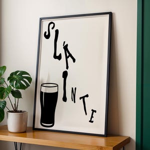 May include: Black framed art print with the word "Sl&aacute;inte" in a stylized font, accompanied by a black silhouette of a pint glass. The print is set against a cream background.