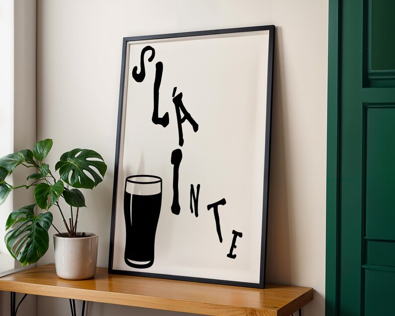 May include: Black framed art print with the word "Sl&aacute;inte" in a stylized font, accompanied by a black silhouette of a pint glass. The print is set against a cream background.