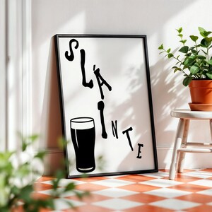 May include: Black framed art print with the word "Sl&aacute;inte" and a pint glass illustration. The artwork is leaning against a white wall, next to a small white stool with a potted plant. The floor has a red and white checkered pattern.