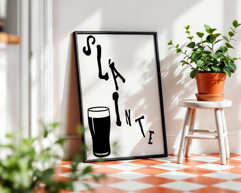 May include: Black framed art print with the word "Sl&aacute;inte" and a pint glass illustration. The artwork is leaning against a white wall, next to a small white stool with a potted plant. The floor has a red and white checkered pattern.