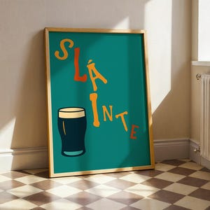 May include: A framed teal poster with the word "SL&Aacute;INTE" in orange letters and a black pint glass illustration. The poster is in a wooden frame and leans against a wall. The floor has a checkered pattern.
