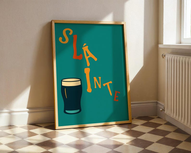 May include: A framed teal poster with the word "SL&Aacute;INTE" in orange letters and a black pint glass illustration. The poster is in a wooden frame and leans against a wall. The floor has a checkered pattern.
