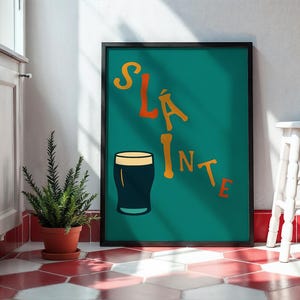 May include: A framed teal poster with the word "SL&Aacute;INTE" in orange and yellow lettering, and a dark pint glass illustration. The poster is leaning against a white wall, next to a small white stool and a potted plant.