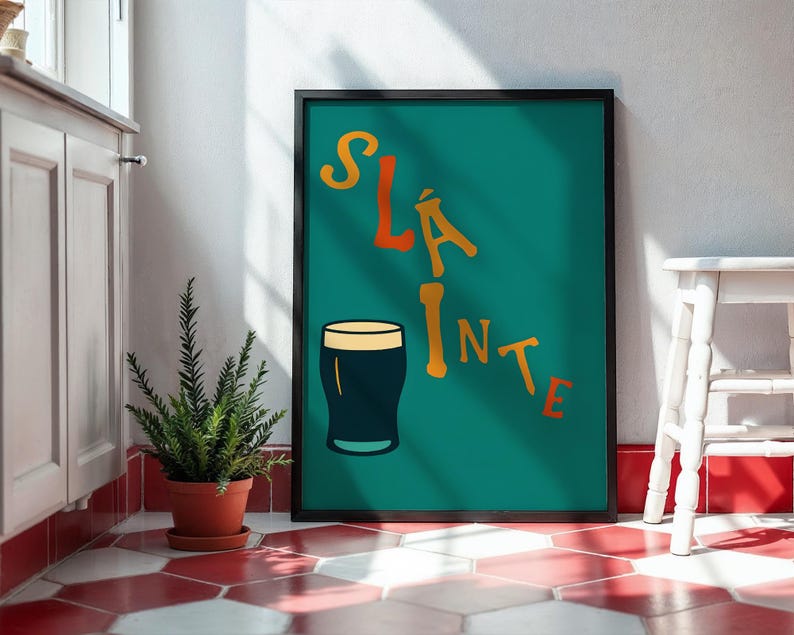 May include: A framed teal poster with the word "SL&Aacute;INTE" in orange and yellow lettering, and a dark pint glass illustration. The poster is leaning against a white wall, next to a small white stool and a potted plant.