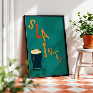 May include: Framed teal poster with the word "Sl&aacute;inte" in orange letters and a black pint of beer illustration. The poster is leaning against a white wall next to a small white stool with a potted plant.