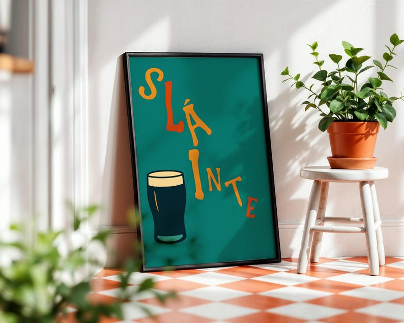 May include: Framed teal poster with the word "Sl&aacute;inte" in orange letters and a black pint of beer illustration. The poster is leaning against a white wall next to a small white stool with a potted plant.