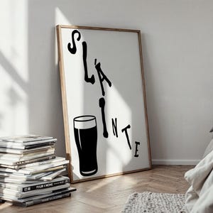 May include: A framed art print with the word "Sl&aacute;inte" in black, along with a black pint glass illustration. The artwork is displayed against a white background, with a wooden frame. The print leans against a wall, next to a stack of magazines.