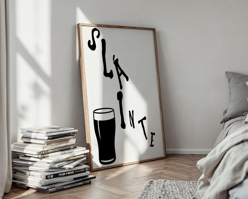May include: A framed art print with the word "Sl&aacute;inte" in black, along with a black pint glass illustration. The artwork is displayed against a white background, with a wooden frame. The print leans against a wall, next to a stack of magazines.