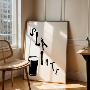 May include: A framed art print with the word "Sl&aacute;inte" in black script, accompanied by a black pint glass illustration. The artwork leans against a wall near a window and a wooden chair.