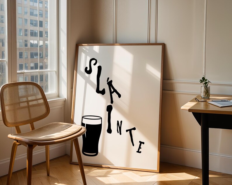 May include: A framed art print with the word "Sl&aacute;inte" in black script, accompanied by a black pint glass illustration. The artwork leans against a wall near a window and a wooden chair.