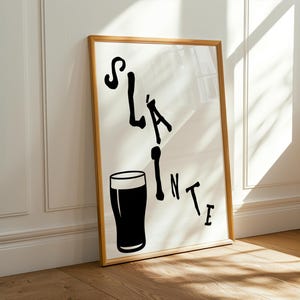 May include: A framed print with the word "Sl&aacute;inte" in black, stylized lettering, accompanied by a black silhouette of a pint glass. The artwork is set against a white background and framed in a light brown wooden frame.