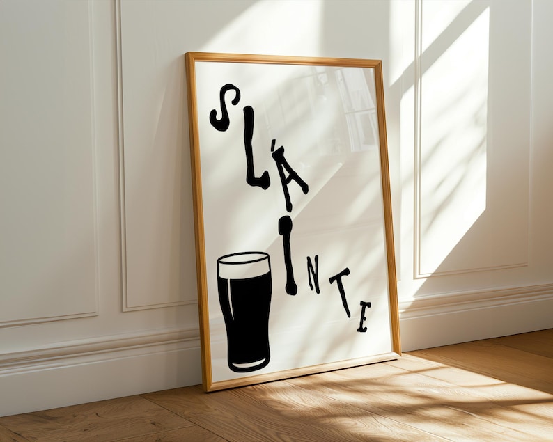 May include: A framed print with the word "Sl&aacute;inte" in black, stylized lettering, accompanied by a black silhouette of a pint glass. The artwork is set against a white background and framed in a light brown wooden frame.