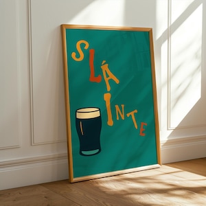 May include: A framed teal poster with the word "Sl&aacute;inte" in orange and yellow lettering, along with a black and white pint glass illustration. The poster is leaning against a white wall and a wooden floor.