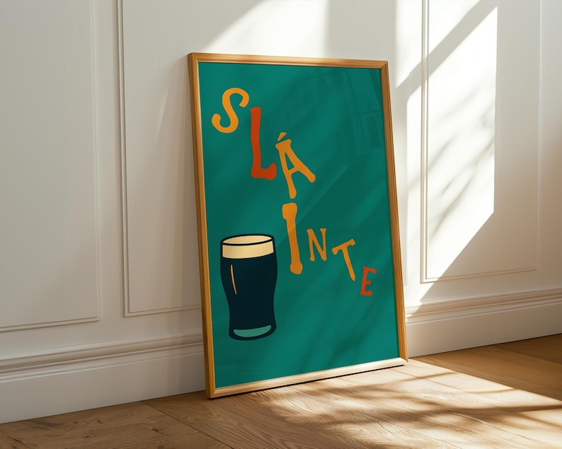 May include: A framed teal poster with the word "Sl&aacute;inte" in orange and yellow lettering, along with a black and white pint glass illustration. The poster is leaning against a white wall and a wooden floor.