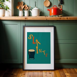 May include: A framed art print with the word "Sl&aacute;inte" in orange and yellow lettering, along with a dark pint glass illustration on a teal background. The print is displayed in a kitchen setting with green cabinets and wooden accents.