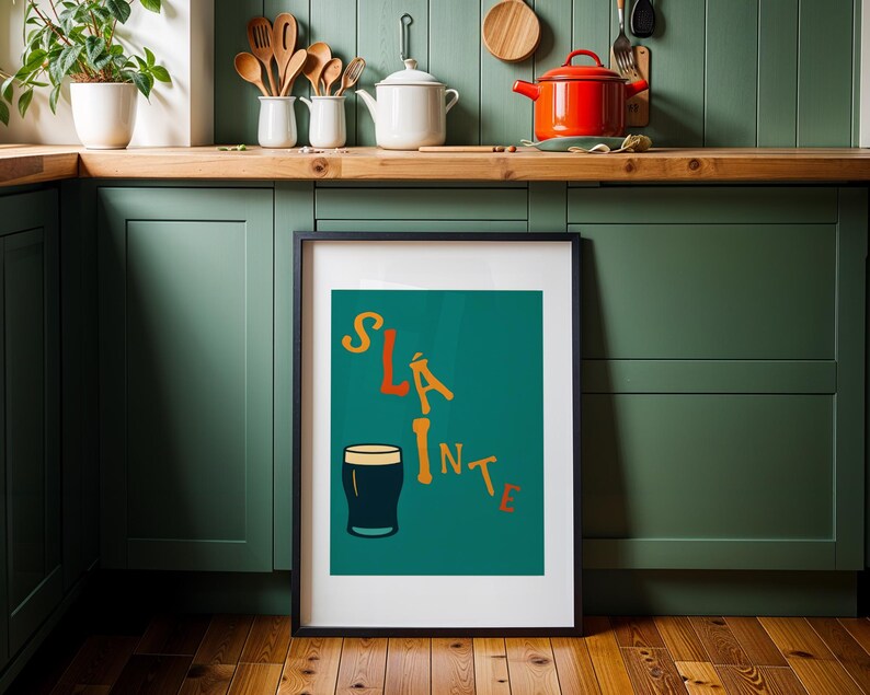 May include: A framed art print with the word "Sl&aacute;inte" in orange and yellow lettering, along with a dark pint glass illustration on a teal background. The print is displayed in a kitchen setting with green cabinets and wooden accents.