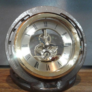 May include: A gold-toned clock with Roman numerals and a visible gear mechanism is set into a gray horseshoe-shaped base.