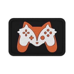 May include: A rectangular embroidered patch with a black border featuring a stylised orange and white fox head design. The fox face is integrated with the shape of a video game controller, incorporating buttons and a directional pad into the white area.