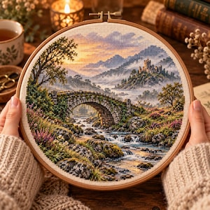 以下が含まれることがあります： 円形の刺繍枠に収められたクロスステッチの風景画。石造りの橋が架かる小川、遠くの丘に建つ城、夕焼けの空が描かれており、岩場の岸辺、緑の木々、色とりどりの野花が細かく表現されています。