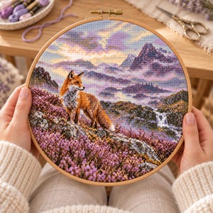 May include: A circular wooden embroidery hoop containing a cross-stitch project depicting a red fox standing on a hillside covered in purple heather. The background features a mountain range under a soft, pastel-colored sky with clouds.