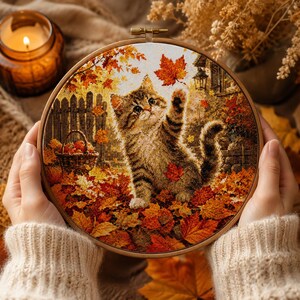 May include: A circular embroidery hoop containing a cross-stitch design of a tabby kitten playing with falling orange and red autumn leaves. The scene includes a wooden fence, a basket of apples, and a lantern in a warm, seasonal setting.