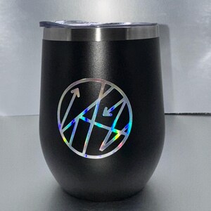 May include: A black, stemless wine tumbler with a silver rim and a clear lid. The tumbler features a circular, holographic design with arrows and geometric shapes. The tumbler is designed to hold approximately 12 ounces.