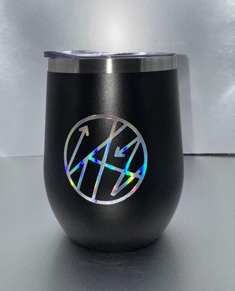May include: A black, stemless wine tumbler with a silver rim and a clear lid. The tumbler features a circular, holographic design with arrows and geometric shapes. The tumbler is designed to hold approximately 12 ounces.