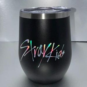 May include: Black insulated tumbler with a silver rim and a clear lid. The tumbler features the iridescent text "Stray Kids" in a stylized font. The tumbler is designed to keep beverages cold or hot.
