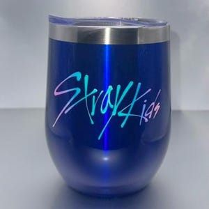 May include: A navy blue insulated tumbler with a silver rim and lid. The tumbler features the text "Stray Kids" in a gradient of pink, blue, and green. The tumbler has a curved, stemless wine glass shape.